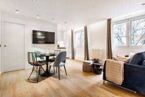 Un salon moderne comprenant un coin repas, un téléviseur à écran plat fixé au mur et une grande fenêtre donnant sur des immeubles.