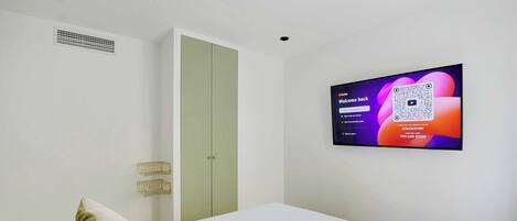 Habitación de hotel con cama, televisor fijado a la pared mostrando una pantalla de bienvenida, una puerta y un pequeño objeto decorativo en la pared.