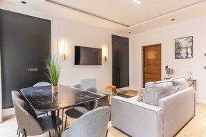 A modern living room with a glass dining table, grey sofas, and a flat-screen TV mounted on the wall.