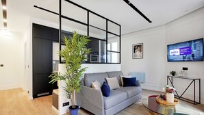 Een moderne woonkamer met een grijze bank, een glazen salontafel, een potplant en een flatscreen televisie aan de wand.