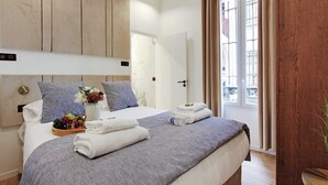 Hotelkamer met een bed, een houten hoofdeinde, een raam met gordijnen en een tafeltje met handdoeken en fruit.