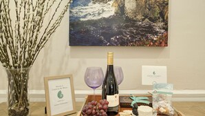 Una mesa de madera con una botella de vino, una copa, un cartel de bienvenida enmarcado, un florero con ramas y una caja de bombones.