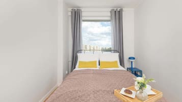 Een slaapkamer met een bed, een nachtkastje, een dienblad met taart en bloemen, en een raam met gordijnen.