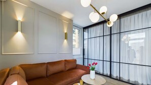 Una sala moderna con un sofá de cuero marrón, una mesita redonda con un jarrón de flores rojas y un ventanal con cortinas translúcidas.