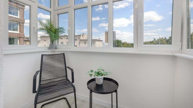 Una habitación con un ventanal grande, una silla negra, una mesita con una planta y vistas a edificios.