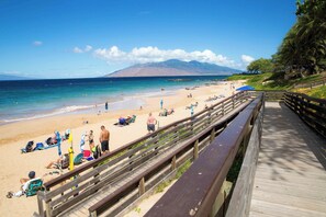 Une promenade en bois qui mène à une plage, avec un ciel dégagé et des montagnes en arrière-plan.