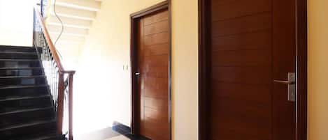 Un pasillo con dos puertas de madera, una escalera con barandillas negras y un piso de tonos claros.