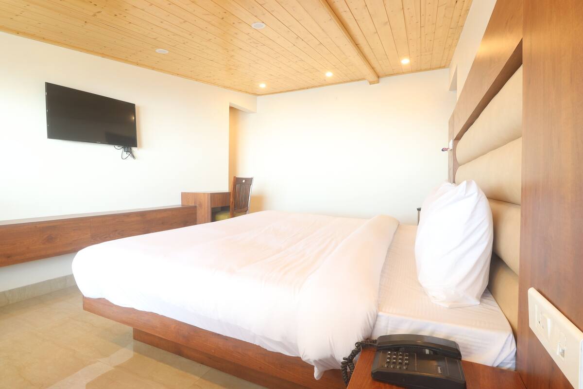 A bedroom with a bed, a telephone, a television mounted on the wall, and a wooden ceiling.