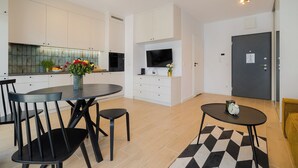 Une cuisine moderne avec des meubles blancs, une table à manger avec des chaises, un téléviseur fixé au-dessus du comptoir et une petite table avec une horloge.