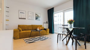 Un salon moderne avec un canapé de couleur moutarde, une table basse et un coin repas comprenant une table ronde et des chaises. Des tableaux encadrés ornent le mur, et de grandes fenêtres offrent une vue sur l’extérieur.