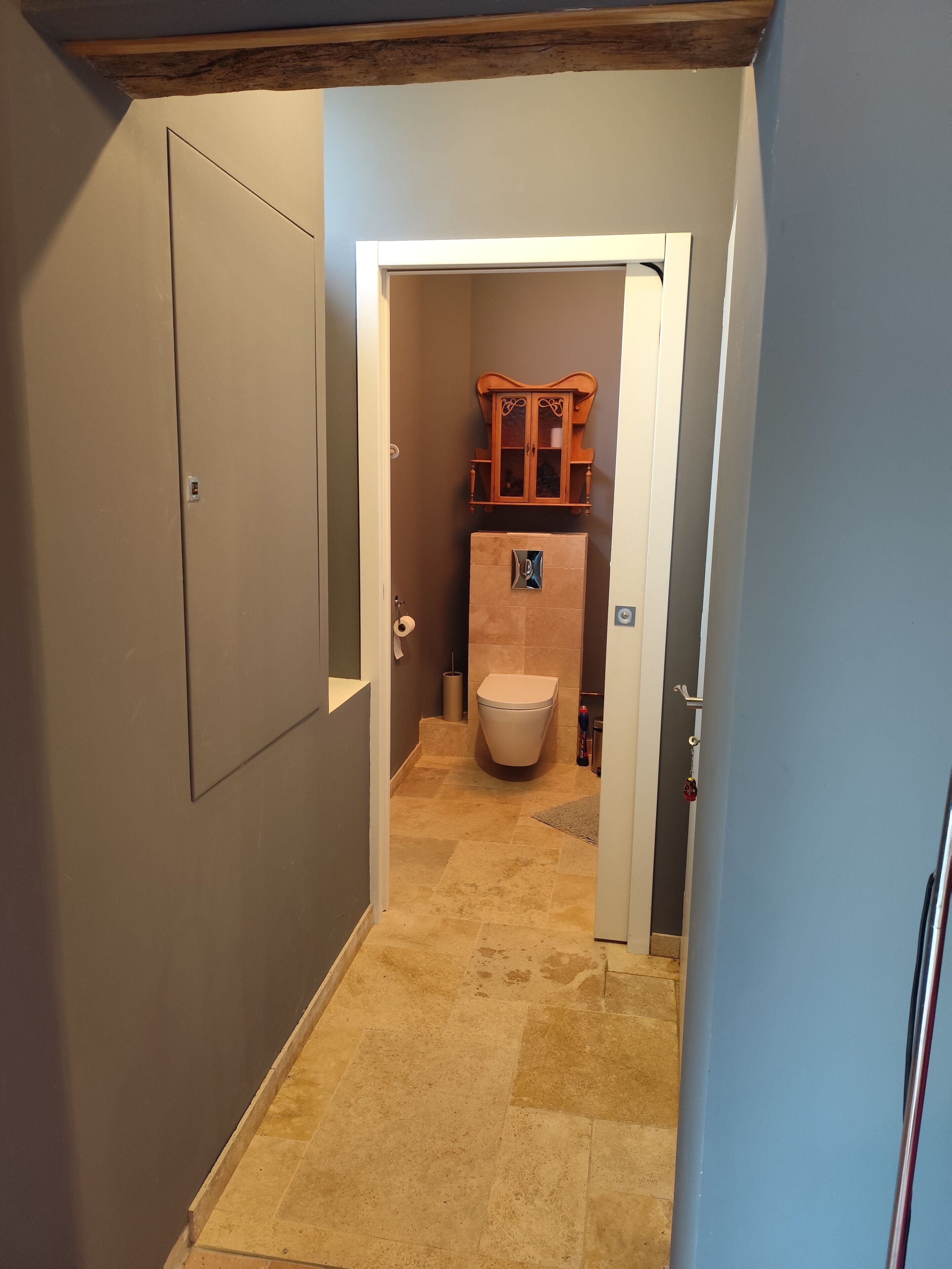 Un couloir étroit mène à une salle de bain équipée de toilettes, d’un meuble et d’un sol carrelé.