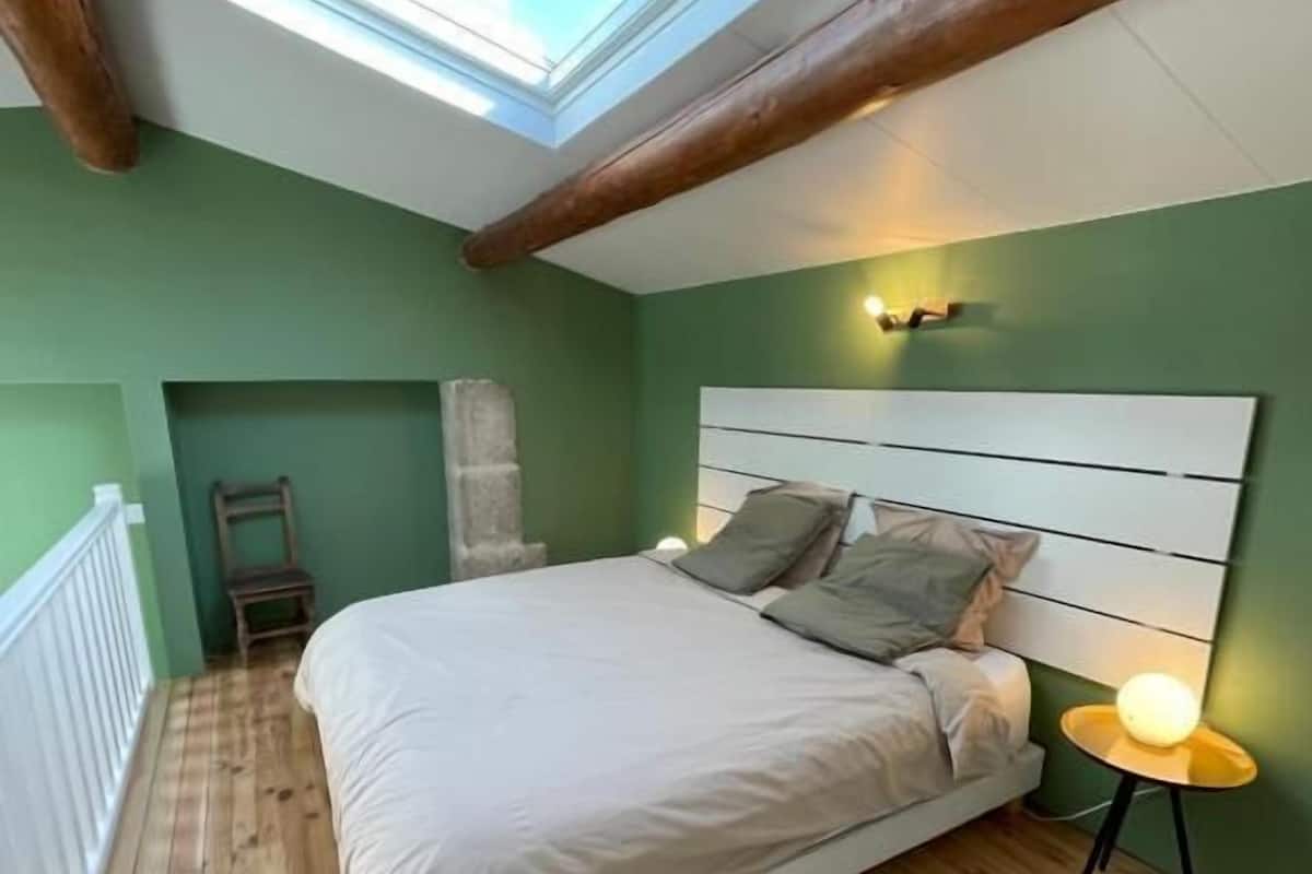 A bedroom with a skylight, wooden beams, a bed with pillows, a bedside table with a lamp, and a view of the outside.