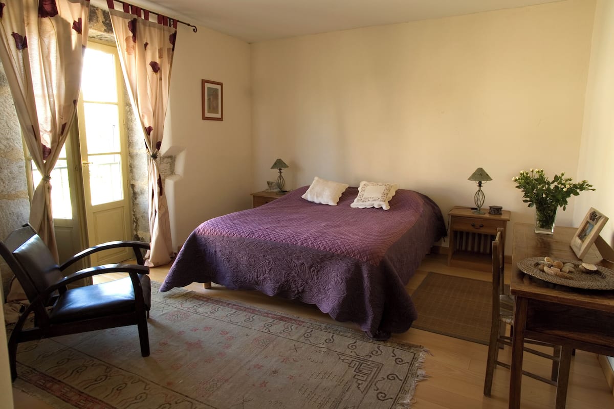 A bedroom with a bed, a chair, a nightstand, a lamp, and a small table with a vase and picture frame.