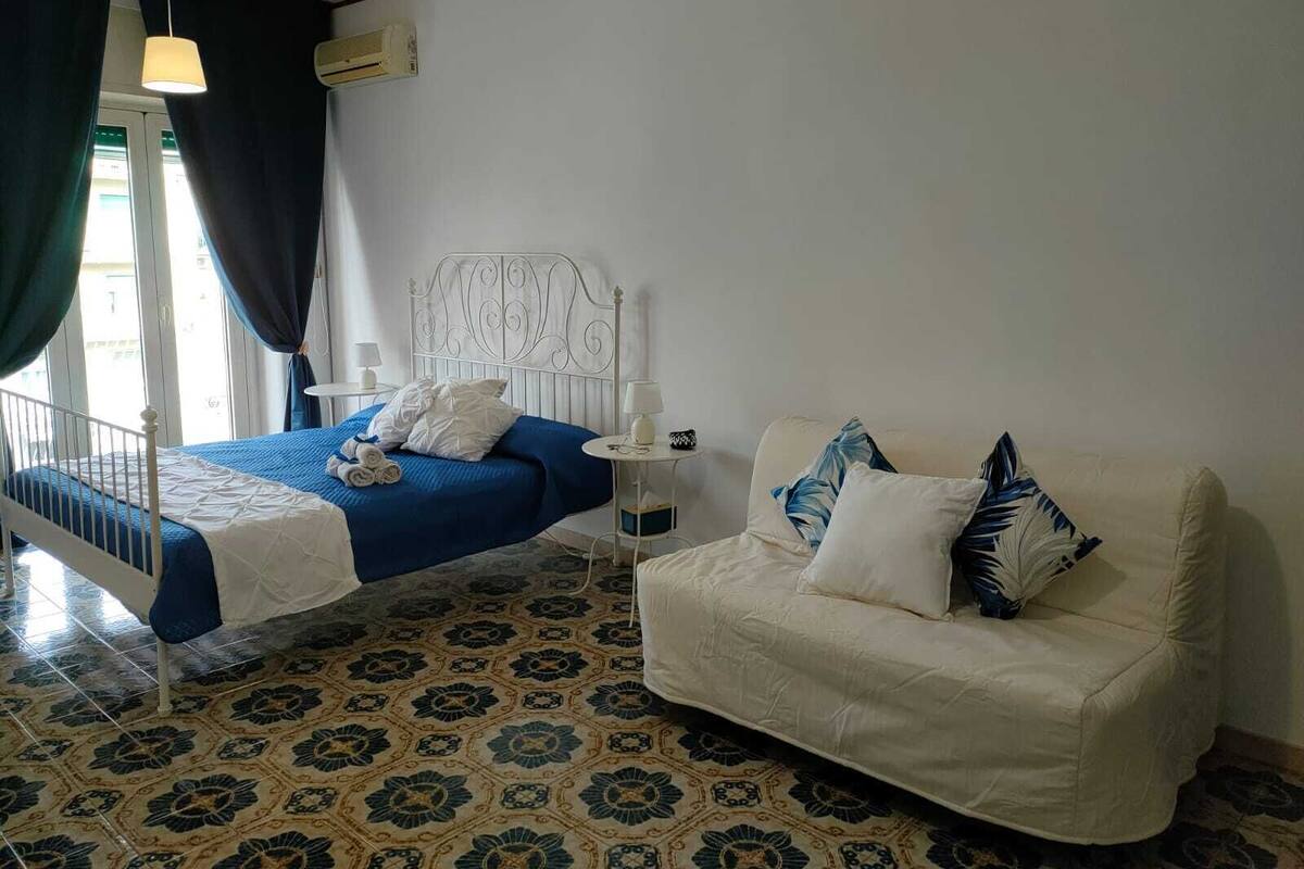 Un dormitorio con cama, sofá blanco, suelo estampado y ventana con cortinas.