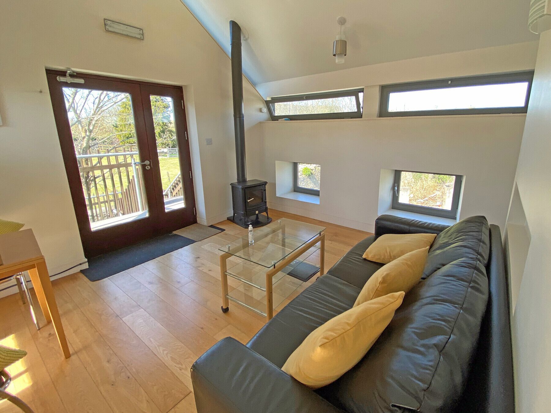 A modern living room with a glass coffee table, a black leather sofa with cushions, a wood-burning stove, and large windows providing natural light.