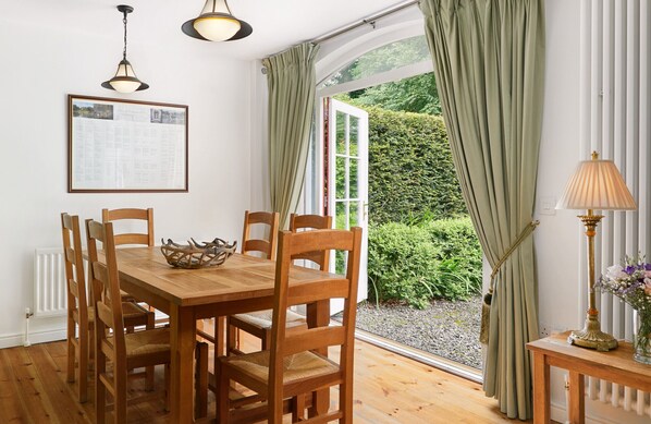 Un espace repas avec une table et des chaises en bois, un menu encadré accroché au mur et une vue sur un jardin, visible à travers une fenêtre ornée de rideaux.