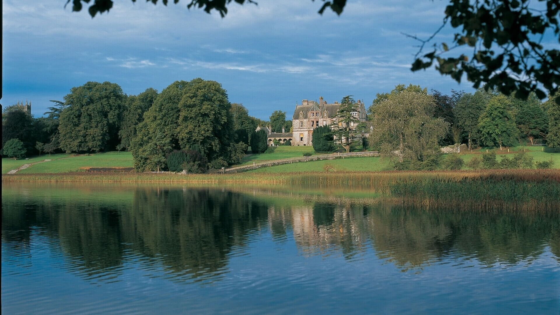 A large, historic building with a symmetrical facade, situated on a lush green landscape with a calm lake reflecting its image.