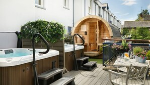 Una bañera de hidromasaje de madera y una sauna de madera en un patio trasero con una terraza, escalones y mobiliario exterior.