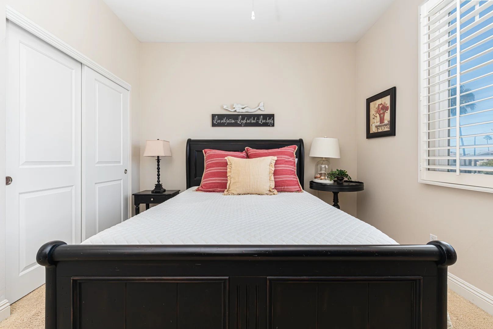 A bedroom with a bed, two bedside tables, a window with blinds, a door, and a wall decoration.