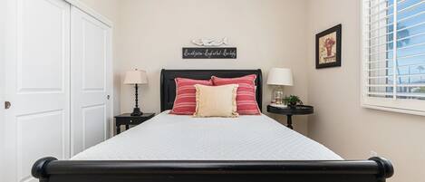 A bedroom with a bed, two bedside tables, a window with blinds, a door, and a wall decoration.