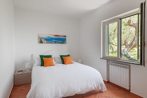 A bedroom with a bed, a nightstand, a lamp, a painting, and a window with a view of trees.