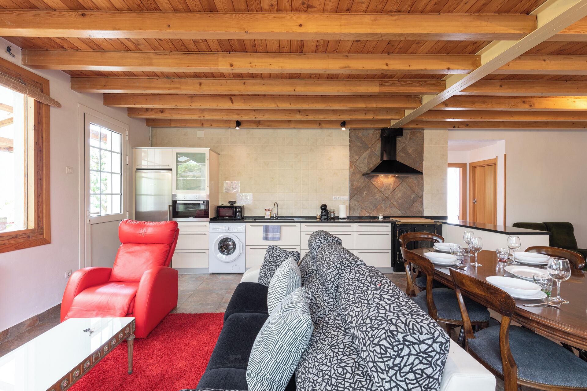 A modern kitchen with wooden beams, a dining area with a long table, and a red leather chair.