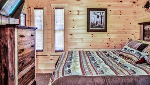 Une chambre avec des murs en bois, un lit avec une literie à motifs, une commode et un téléviseur posé sur un meuble.