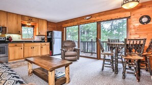 Ein Blockhaus-Interieur mit einem Holzespeisetisch, Stühlen, einer Couch, einer Küche mit Holzschränken, einem Kühlschrank und einem großen Fenster mit Blick auf Bäume.