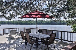 Una terraza con una mesa redonda y sillas, un paraguas rojo y vistas a un lago con árboles al fondo.
