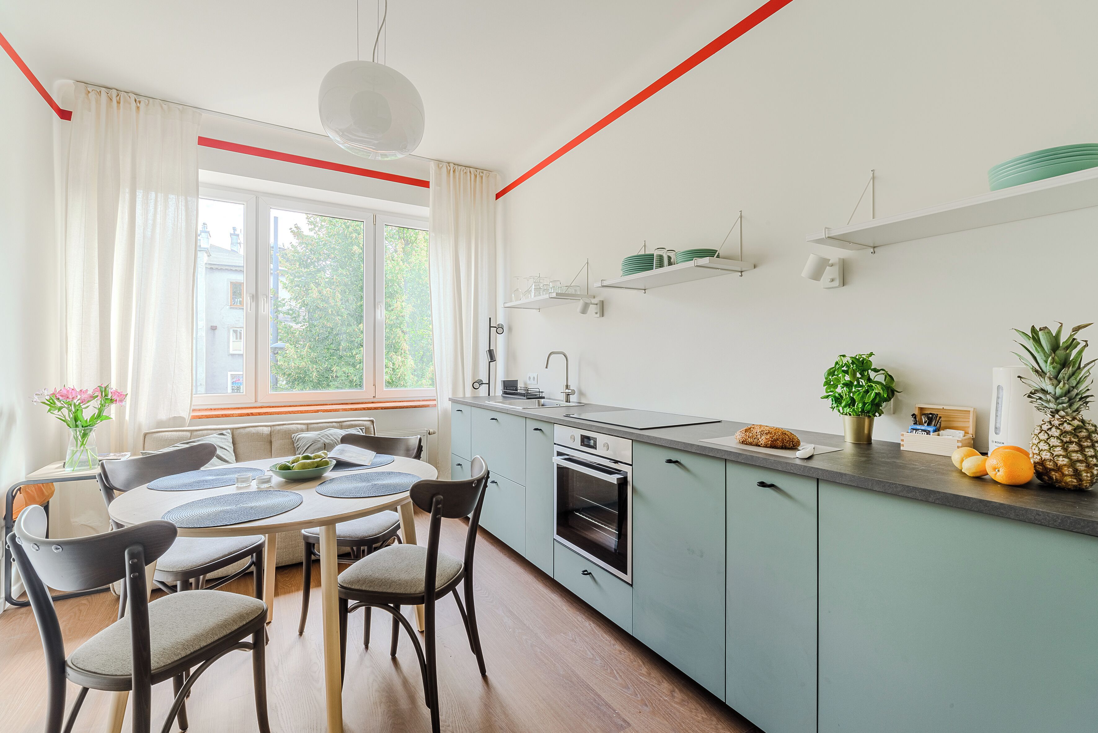 Una cucina moderna con zona pranzo, caratterizzata da un'ampia finestra, mobili chiari e un piano di lavoro con diversi oggetti.