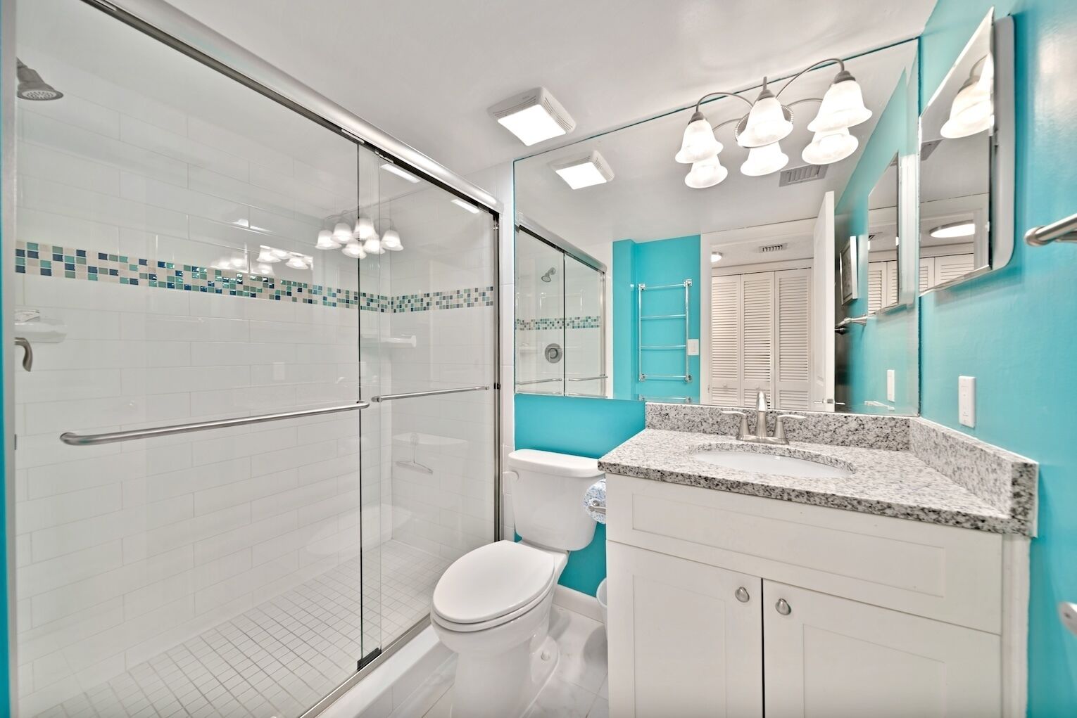 A modern bathroom with a glass shower enclosure, a white toilet, a turquoise accent wall, and a granite countertop sink.