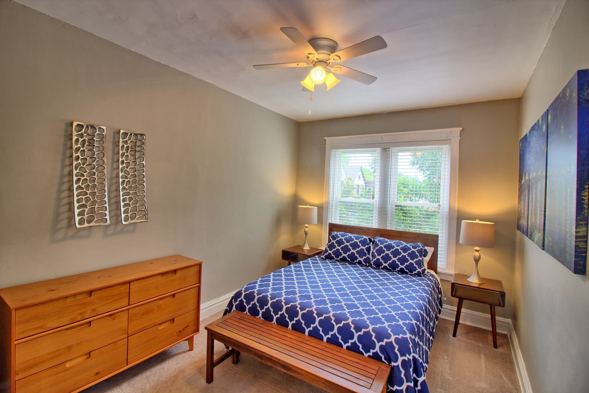 A bedroom with a bed, wooden dresser, two nightstands, a ceiling fan, and a window with blinds.
