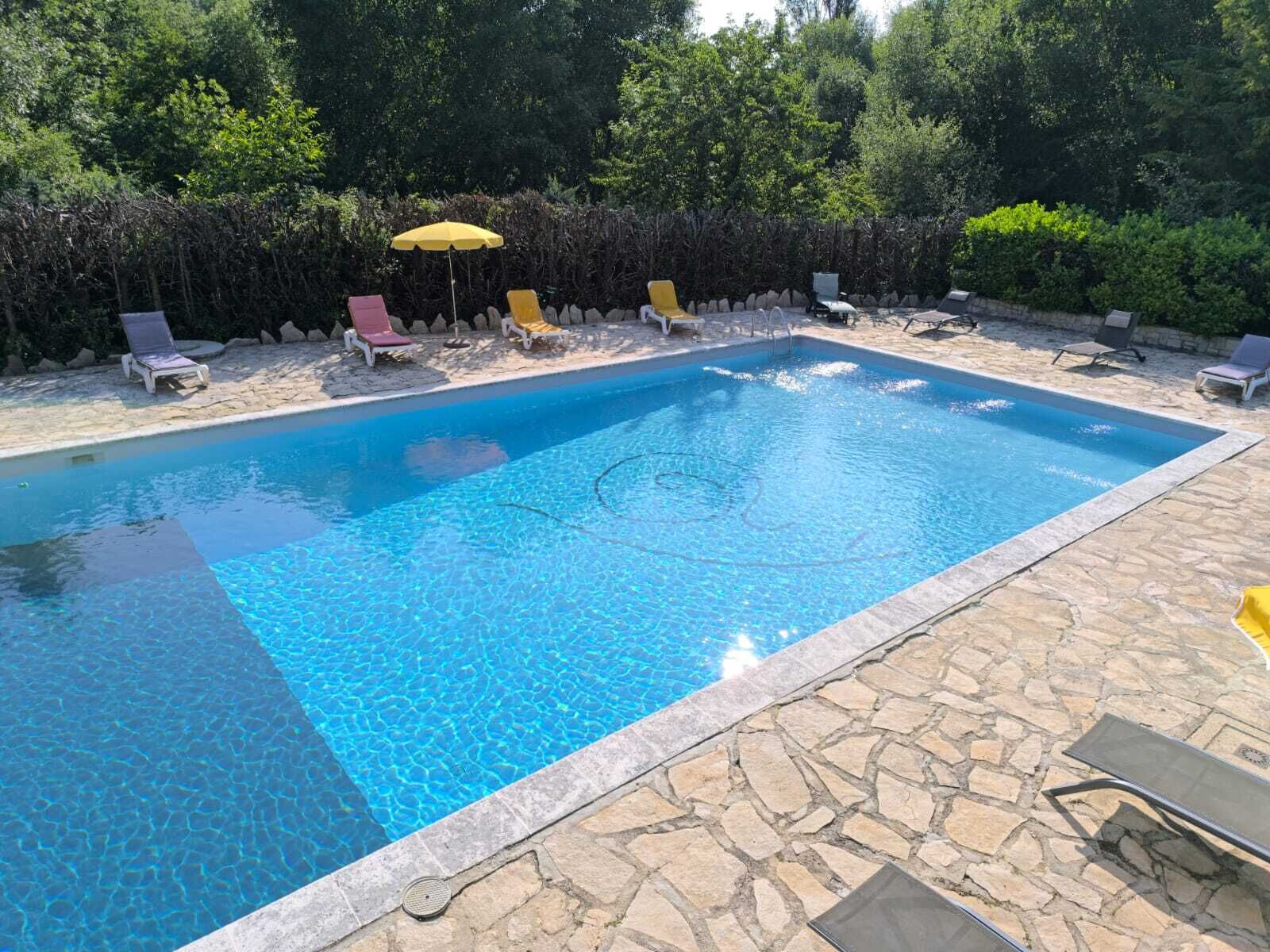 A rectangular swimming pool with clear blue water, surrounded by stone tiles and equipped with several lounge chairs and a yellow umbrella.