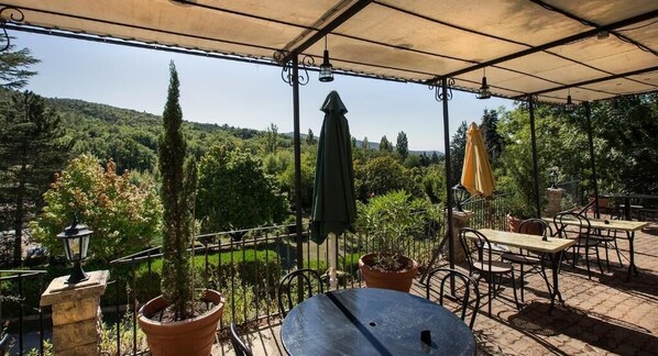 An outdoor patio with tables and chairs, surrounded by potted plants and a view of a forested hillside.