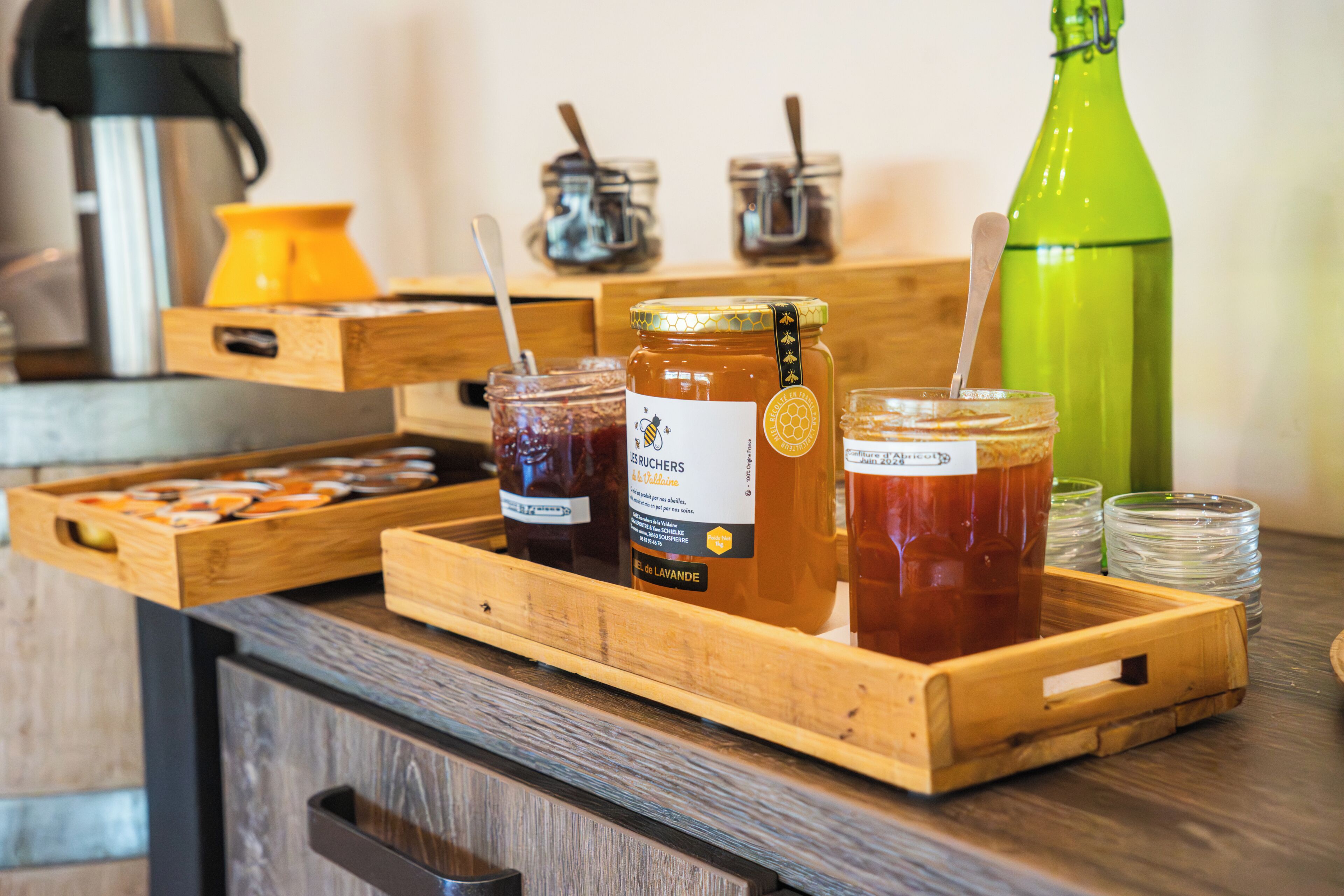 A wooden tray with jars of honey and a green bottle on a counter.