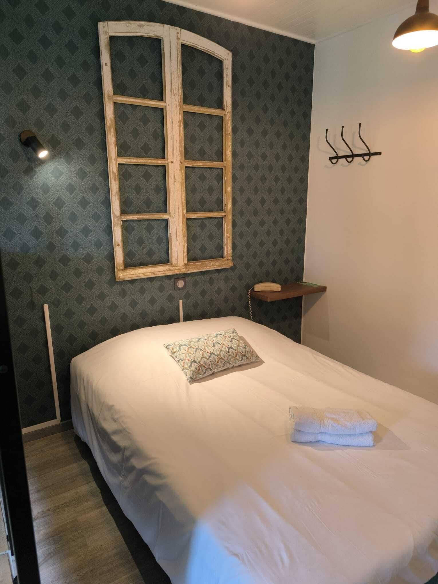 Una cama bien hecha con una almohada estampada y una toalla doblada, una ventana con marco de madera y un gancho fijado a la pared.