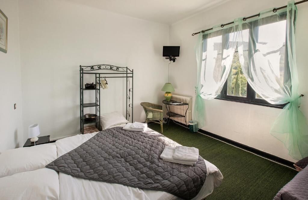 Un dormitorio con cama, mesita de noche, silla, radiador, televisor y ventana con cortinas traslúcidas.