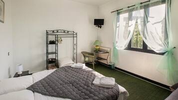 Un dormitorio con cama, mesita de noche, silla, radiador, televisor y ventana con cortinas traslúcidas.