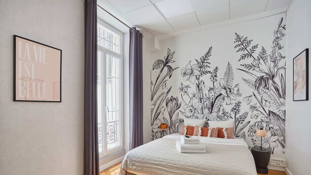 Une chambre avec une grande fenêtre, un lit recouvert de draps blancs, un papier peint à motifs floraux et un tableau encadré accroché au mur.