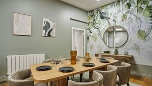 Un coin repas avec une table en bois dressée pour quatre personnes, entourée de chaises rembourrées. Un radiateur est visible sur la gauche, et un grand miroir est accroché au mur.