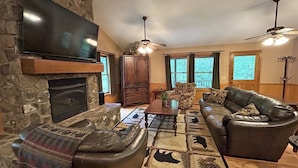 A cozy living room with a stone fireplace, leather sofa, and a flat-screen TV mounted on the wall. - Dream on a Stream Log Cabin Lodge That Has It All! (Almond)