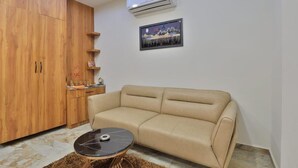 A living room with a beige sofa, a black coffee table, wooden cabinets, and an air conditioning unit. - Hotel TGR Rooms (Palanpur)