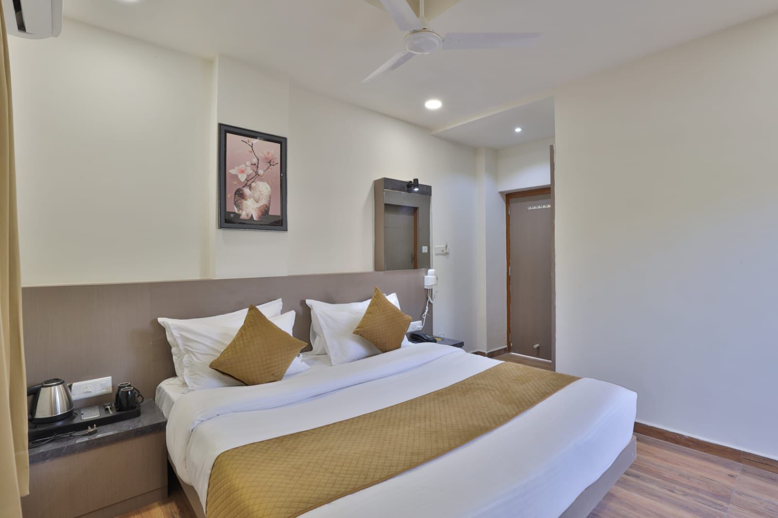A hotel room with a large bed, a nightstand, a coffee maker, a framed picture on the wall, and a door in the background.