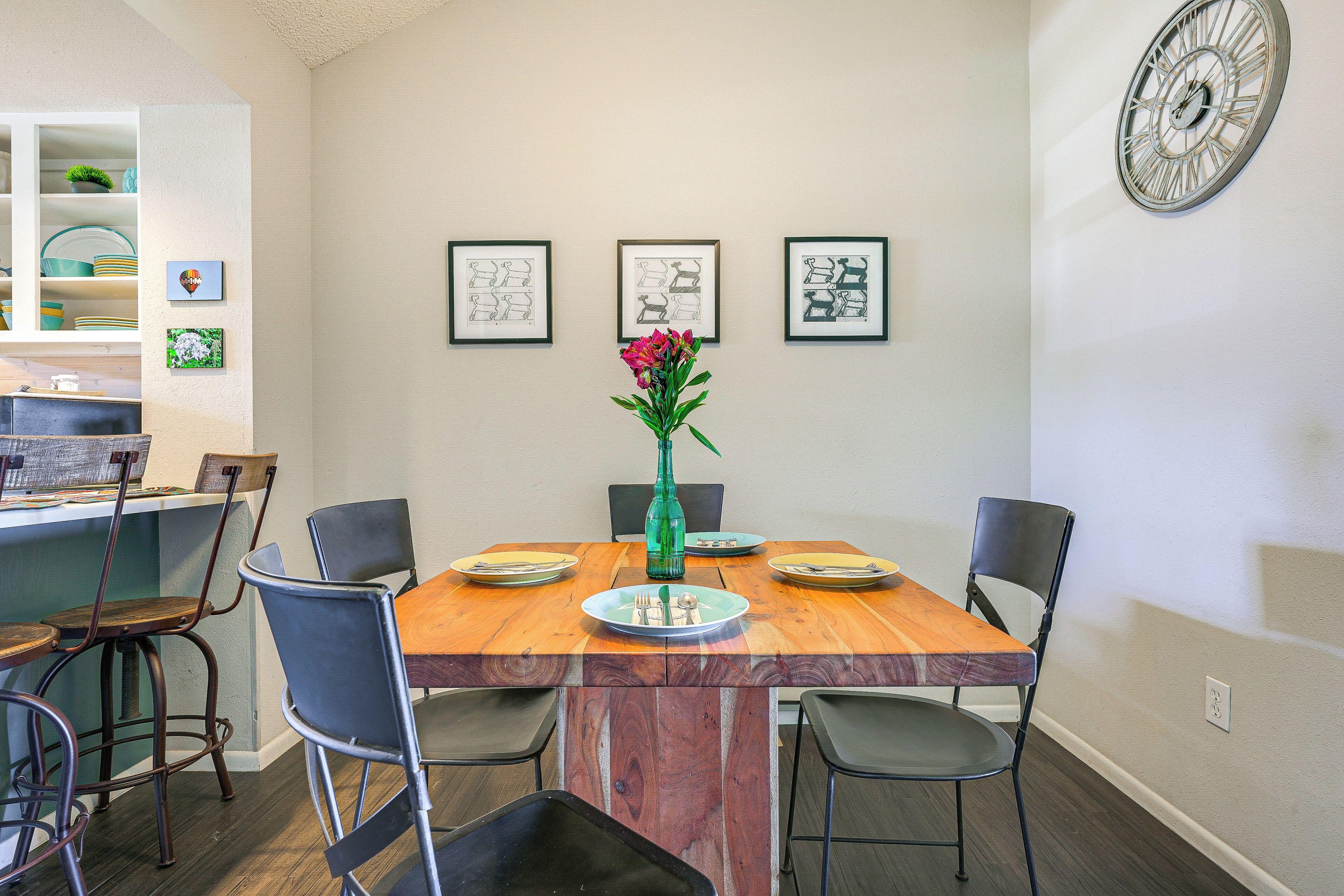 A dining area with a wooden table, four chairs, and a vase with flowers. There are framed pictures on the wall and a clock.