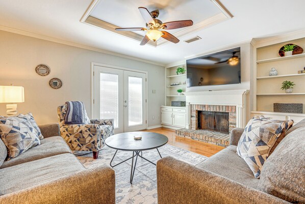 A living room with a fireplace, a ceiling fan, and a large TV. - Mobile Townhome Near University of South Alabama! (Mobile)