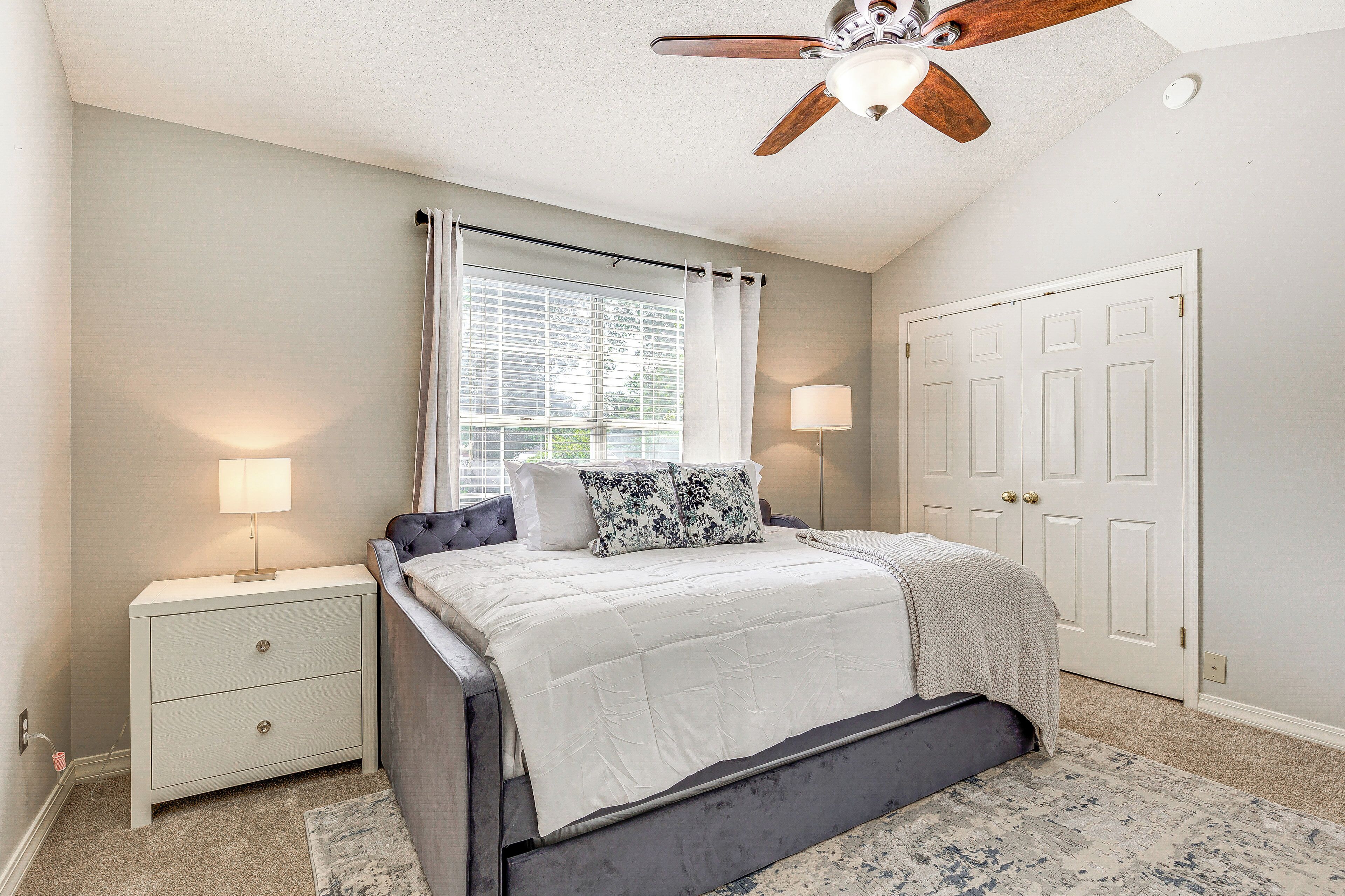 A bedroom with a bed, nightstands, a ceiling fan, a window with curtains, and a door.