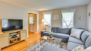 A living room with a flat-screen TV on a wooden TV stand, a grey sofa, a glass coffee table, and a patterned rug.
