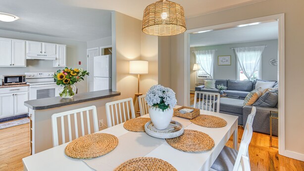 A modern kitchen with white cabinets, a microwave, and a stove. There is a dining area with a white table, woven placemats, and a vase of flowers.