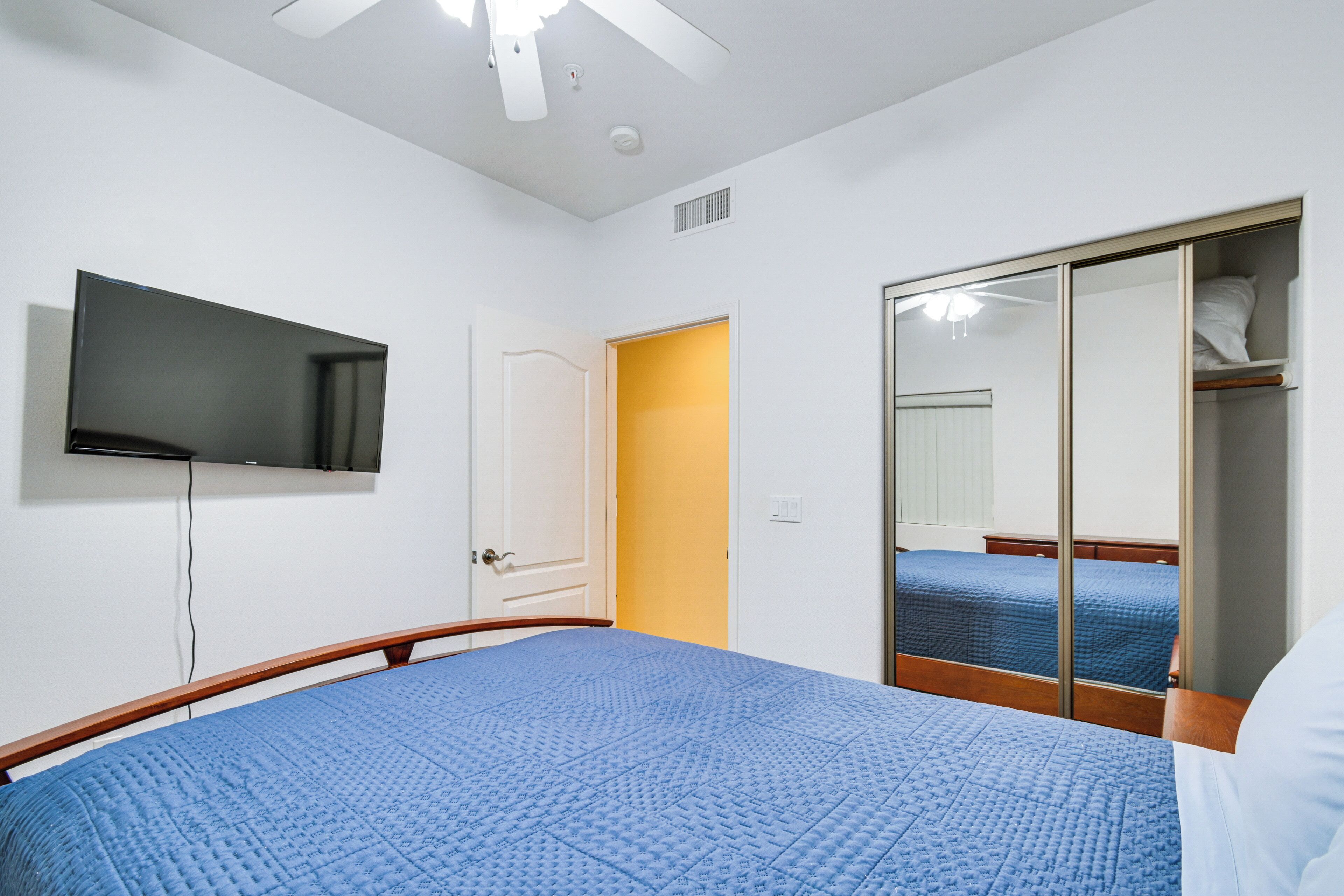 Une chambre à coucher comprenant un lit, un téléviseur fixé au mur, une armoire avec un miroir et une porte donnant sur une autre pièce.