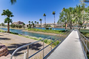 A riverside walkway with a metal railing, palm trees, and a building in the background.